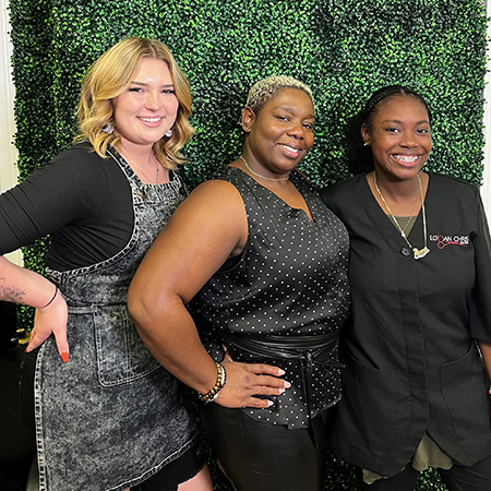 Three smiling women in front of an artificial green foliage backdrop